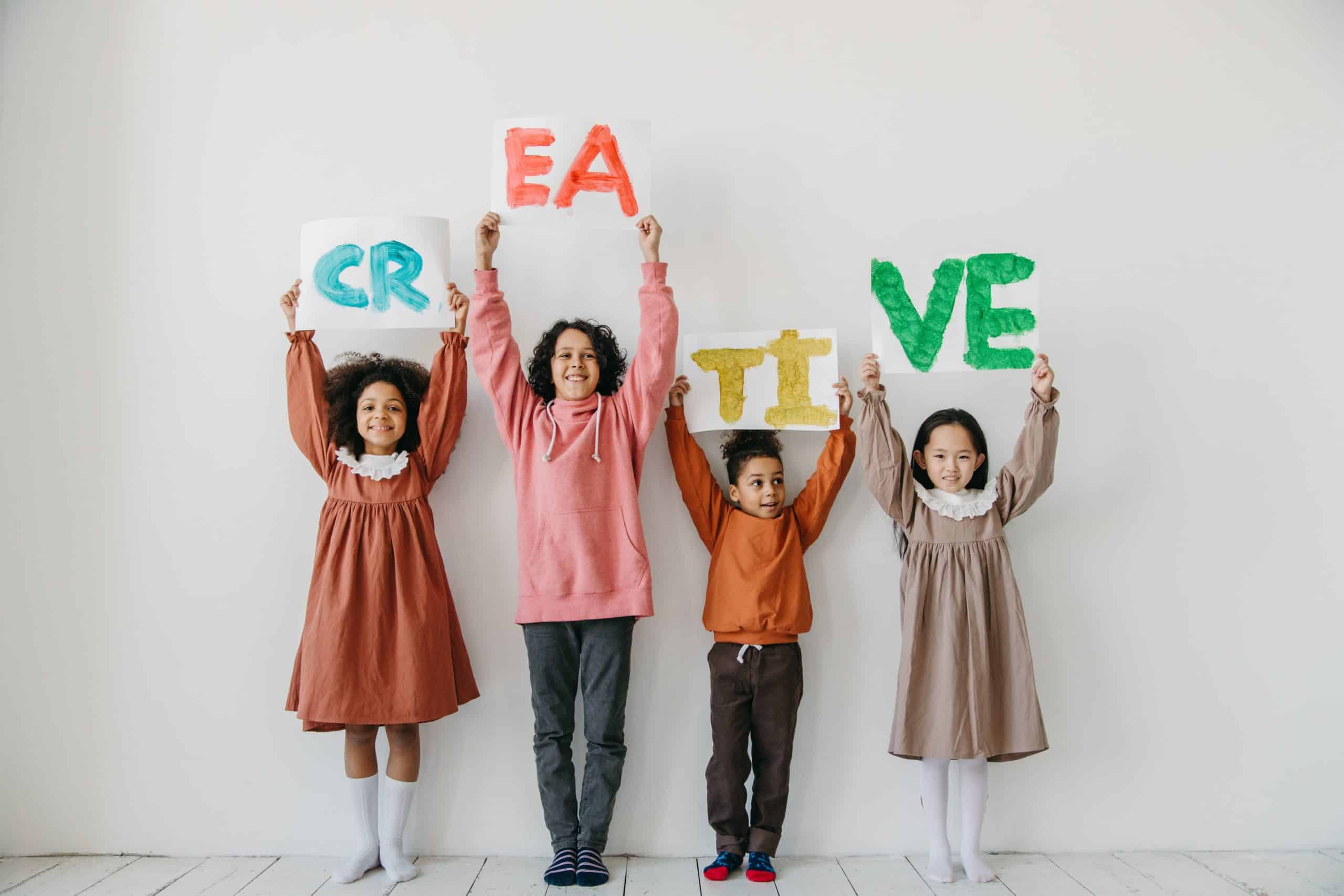 About 1 Four children and one person hold colorful signs spelling "CREATIVE" against a plain white background. They smile, displaying diversity and joy.