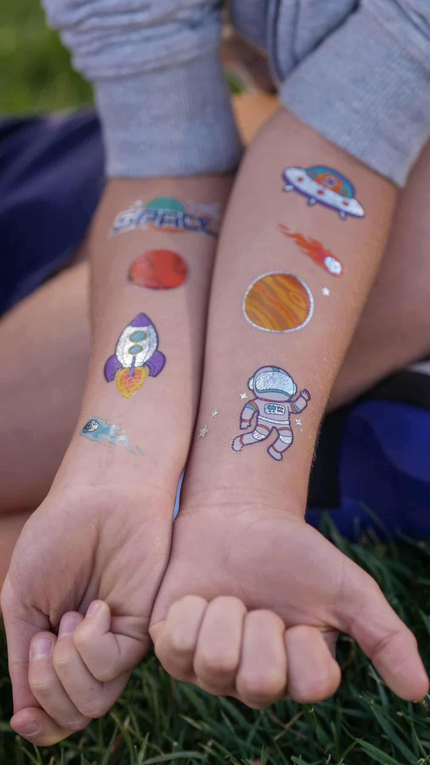 About 6 Two arms with colorful space-themed temporary tattoos, featuring rockets, planets, and an astronaut, rest on grass.
