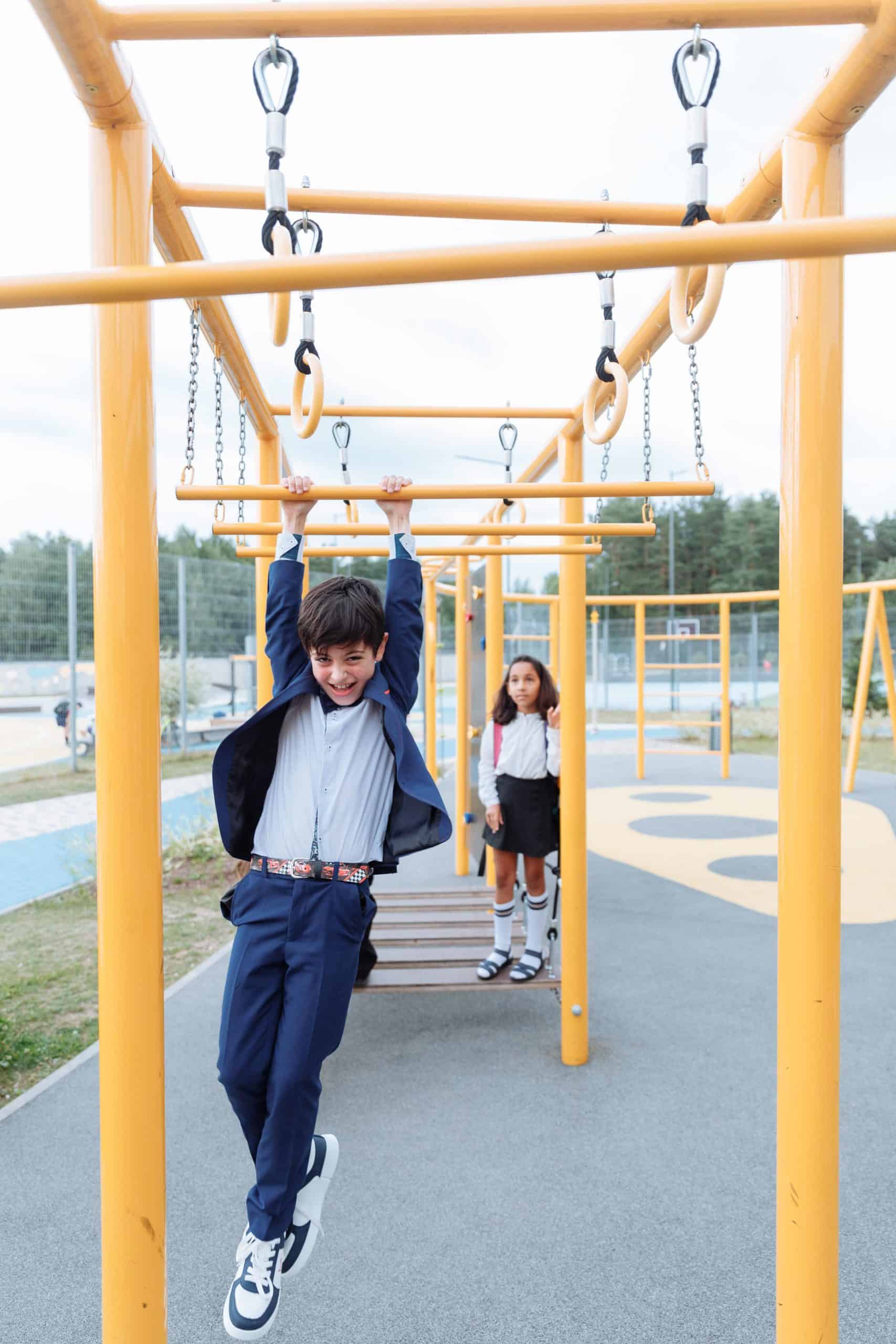 About 3 A child in a blue suit hangs on yellow monkey bars at a playground, with another child standing nearby, surrounded by trees.